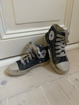 Svarta Converse Chuck Taylor High - Klassiska svarta Converse Chuck Taylor All Star high-top sneakers med vita gummisulor och tåhättor. Skorna har vita snören och tydliga kontrastsömmar samt den ikoniska loggan på sidan. Ovandelen är i canvas och sulan i gummi.