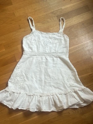 Vit sommarklänning - Söt vit kortklänning med tunna axelband och volangkant nertill. Klänningen har fina broderade blommor och spetsdetaljer i midjan och upptill. Perfekt för varma dagar och har en lätt och luftig känsla. Storlek M men passar även S
