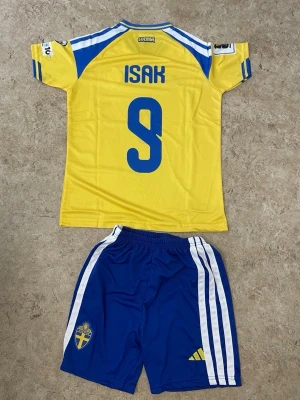 Sverige matchställ Isak #9 Adidas - Säljer ett officiellt Sverige matchställ från Adidas med Isak och nummer 9 på ryggen. Tröjan är gul med blå detaljer och korta ärmar, byxorna är blå med vita ränder och Sveriges landslagsemblem. Perfekt för fotbollsträning eller match.