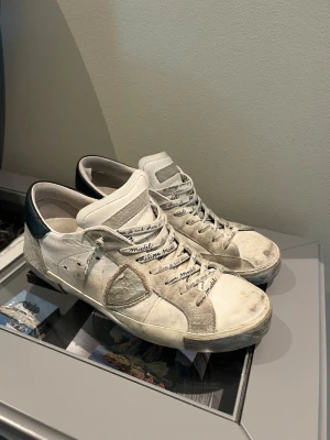 Philippe Model vita sneakers - Snygga vita sneakers från Philippe Model med grå mockadetaljer och svart hälparti. Skorna har klassisk låg profil, perforerade sidor och vita snören med logotyptext. Ovandelen är i läder och mocka för en trendig och avslappnad look.