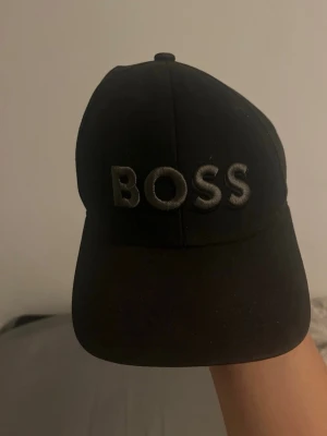 Svart keps från BOSS med brodyr - Säljer en svart keps från BOSS med tydlig broderad logga framtill. Klassisk böjd skärm och justerbar rem bak. Tillverkad i bomullsmix för en bekväm passform. Perfekt för dig som vill ha en stilren och enkel accessoar.