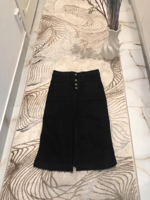 Svart långkjol med knappar - Snygg svart långkjol med raka linjer och fyra silverfärgade knappar framtill. Kjolen har två stora fickor fram och en liten slits nedtill. Perfekt till en cool vardagslook och funkar med både sneakers och boots.