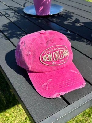 Rosa keps med New Orleans-brodyr - Snygg rosa keps med broderad patch där det står 'New Orleans' och 'Let the good times roll'. Kepsen har en sliten, vintage-look med flera slitningar och fransar på skärmen. Tillverkad i bomullstyg och justerbar baktill.
