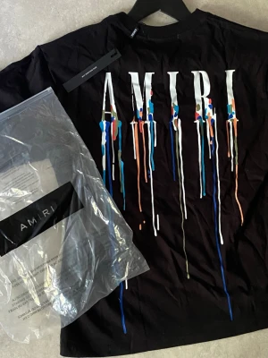 Svart AMIRI t-shirt med färgstänk - Cool svart t-shirt från AMIRI med stort tryck på ryggen och mindre tryck på bröstet. Trycket består av vita bokstäver och färgstänk i blått, orange, rött och grönt som rinner ner. Klassisk passform och rund hals, tillverkad i mjuk bomull.