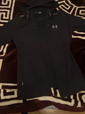 Svart hoodie från Under Armour - Snygg svart hoodie från Under Armour med dragkedja framtill och diskret logga på bröstet. Modellen har huva och är tillverkad i ett mjukt, stretchigt material som passar perfekt för en sportig look. Praktisk ficka med dragkedja på sidan.