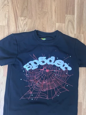 Svart Sp5der t-shirt med spindelnätsmotiv - Cool svart t-shirt med tryck av ett rött spindelnät och texten 'sp5der' i stora bokstäver på bröstet. T-shirten har korta ärmar och en klassisk rund halsringning. Perfekt för dig som gillar streetwear och grafiska prints.
