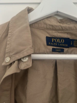 Beige skjorta från Polo Ralph Lauren - Klassisk beige skjorta från Polo Ralph Lauren i slim fit-modell. Skjortan har lång ärm, knappar framtill och ikonisk broderad logga på bröstet. passar perfekt till en avslappnad men stilren look. Kan mötas i Västerås 