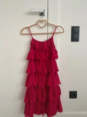 Rosa volangklänning med smala axelband - Supersöt rosa klänning med flera lager volanger och smala axelband. Klänningen har en luftig och flowy känsla, perfekt för sommarens alla tillfällen. Materialet känns lätt och svalt, och modellen är kort med en feminin siluett.