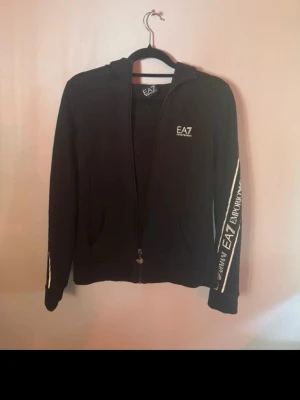Svart hoodie från EA7 Emporio Armani - Svart hoodie med dragkedja från EA7 Emporio Armani. Snygga vita logotexter längs båda ärmarna och liten logga på bröstet. huva och två fickor framtill. Lappen i nacken sitter bara fast i ena änden, gick av när jag hängde upp den, annars fint skick. 