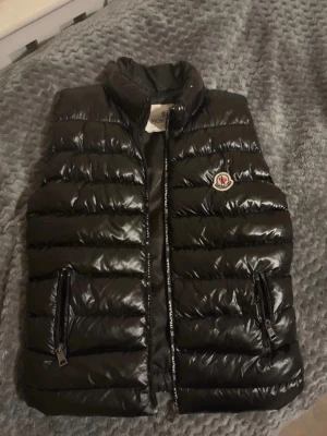Svart dunväst från Moncler - …Går såklart att pruta… Har lite problem me fickorna och ett litet hål. Snygg svart dunväst från Moncler med glansig finish och klassisk logga på bröstet. Västen har två dragkedjeförsedda fickor och hög krage. Perfekt för lager-på-lager och ger en trendig look till din outfit.