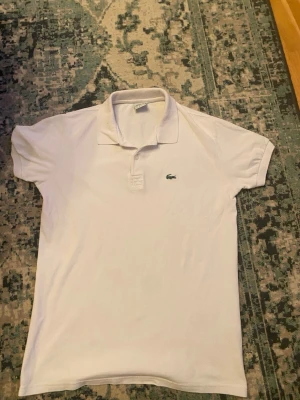 Vit pikétröja från Lacoste - Klassisk vit pikétröja från Lacoste med korta ärmar och krage. Tröjan har den ikoniska gröna krokodilloggan broderad på bröstet och är tillverkad i bomull. Perfekt för en clean och sportig stil.