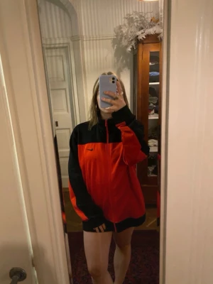 Nike röd och svart zipjacket - Snygg röd och svart jacka från Nike med dragkedja framtill och hög krage. Jackan har en sportig look med svarta detaljer på axlar och ärmar samt diskret Nike-logga på bröstet. Perfekt för dig som gillar streetwear och vill ha en lätt jacka till vardagen.