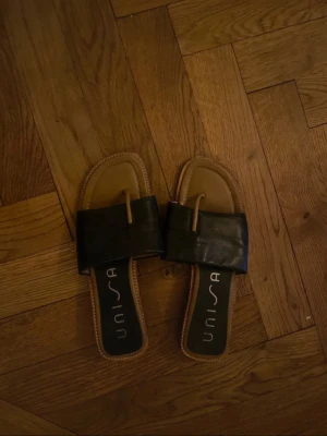 Svarta slip-in sandaler från Unisa - Stilrena svarta slip-in sandaler från Unisa med bred rem i skinn och fyrkantig tå. Sandalerna har låg klack och en brun sula med kontrastsömmar. Perfekta för en avslappnad och trendig look.