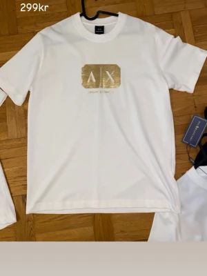 2st T-shirt    Vit t-shirt med gulddetaljer AX & Balmain - Två stilrena vita t-shirts, en från Armani Exchange med guldfärgat AX-tryck och en från Balmain Paris med guldfärgad logga. Båda har klassisk passform, rund hals och korta ärmar. Perfekta för dig som gillar exklusiva märken och enkel design.                  2st för endast  500kr
