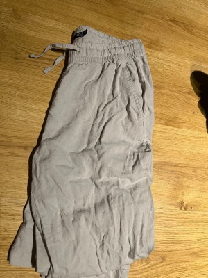 Beige linnebyxor från 157 - Säljer ett par beige linnebyxor från 157 i storlek XS. Byxorna har resårmidja med dragsko och sidofickor. Sköna och luftiga med avslappnad passform, perfekta för sommaren. Materialet är linne vilket ger en sval känsla.