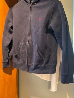 Ralph Lauren Zip-Hoodie - Säljer en äkta marinblå zip-hoodie från Polo Ralph Lauren. Storleken är L (14-16 år). Tröjan är köpt ny på Kidsbrandstore för 1100 kr och är i fint begagnat skick då den knappt använts, men den har tyvärr blivit lite lätt solblekt på framsidan. Äktheten är 100% säkerställd och original-etiketter (inklusive den gula QR-lappen) finns kvar. Hör av dig vid ytterliggare funderingar