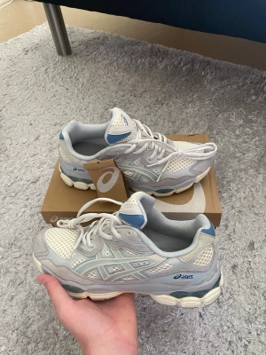 Vita och grå Asics sneakers - Storlek 42,5. Snygga sneakers från Asics i vitt och ljusgrått med blå detaljer. Ovandelen är i mesh och syntetmaterial för en sportig vibe. Klassisk Asics-logga på sidan och tungspetsen. Skorna har en chunky sula och lågt skaft.