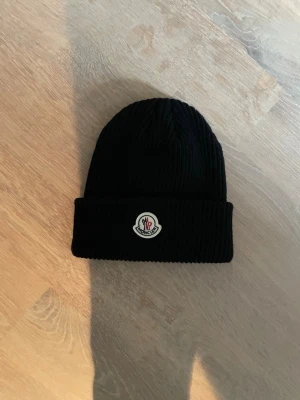 Svart ribbstickad mössa Moncler - Snygg svart ribbstickad mössa från Moncler med uppvikt kant och klassisk Moncler-logga framtill. Mössan är tillverkad i ett mjukt och varmt material som passar perfekt för kyliga dagar. Enkel och stilren design som funkar till det mesta.