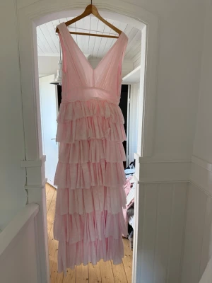 Ljusrosa volangklänning med v-ringning - Säljer en lång, ljusrosa klänning med djup v-ringning och flera lager volanger. Klänningen är ärmlös och har en romantisk look som passar perfekt till speciella tillfällen. Materialet känns lätt och luftigt.