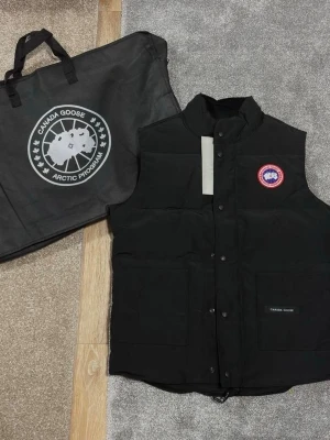 Svart dunväst från Canada Goose - Snygg svart dunväst från Canada Goose med klassisk logotyp på bröstet och praktiska framfickor. Västen har hög krage och stängs med tryckknappar. Perfekt för lager-på-lager och håller dig varm under kyliga dagar.