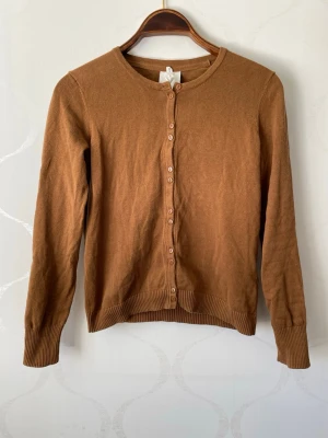 Brun kofta med rund hals - Klassisk brun kofta med rund halsringning och knappar framtill. Långärmad modell med ribbade muddar vid ärmslut och nederkant. Perfekt att slänga över en t-shirt eller linne för en chill look. Koftan har en unik knapp sättning.