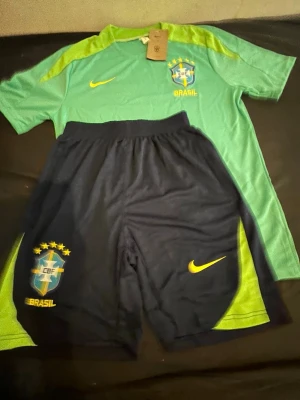 Nike Brasil fotbollskit grön/blå  - Säljer ett Brasilien fotbollskit från Nike i storlek S. Tröjan är ljusgrön med limegröna detaljer och har CBF-logga och Nike-swoosh på bröstet. Shortsen är mörkblå med limegröna inslag, CBF-märke och Nike-logga. Perfekt för träning eller match!