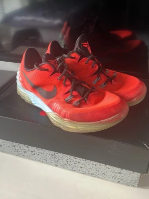  Kobes  - Säljer ett par Nike Kobe basketskor i rött meshmaterial med svart swoosh och detaljer. Skorna har svart snörning, genomskinlig sula och coolt mönstrad häl i blått och vitt. Perfekta för basket eller streetwear och har en låg profil för smidiga rörelser.