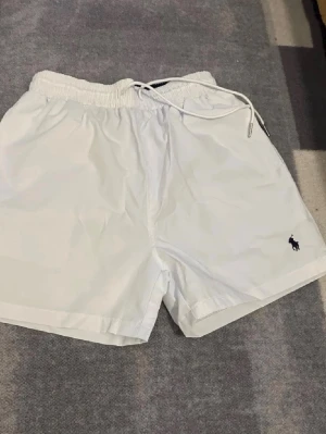 Badshorts - Fräscha vita shorts från Polo Ralph Lauren med elastisk midja och snörning. Klassisk design med liten broderad logga framtill och praktisk bakficka med lock. Perfekta för sommardagar och chill häng.