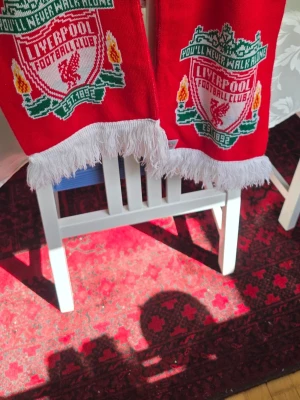 Liverpool FC röd halsduk med tofsar - Röd halsduk med Liverpool FC:s klubbmärke och texten 'You'll Never Walk Alone' i grönt och vitt. Halsduken har vita fransar i ändarna och detaljer i gult och grönt. Perfekt för dig som vill visa ditt stöd för laget.