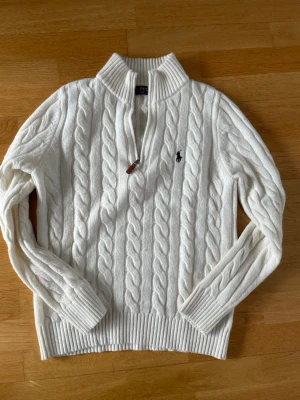 Vit kabelstickad tröja Polo Ralph Lauren - Vit kabelstickad tröja från Polo Ralph Lauren med halv dragkedja och ribbade muddar. Klassisk design med broderad logga på bröstet. Perfekt för lager-på-lager och snygg till jeans eller kjol.