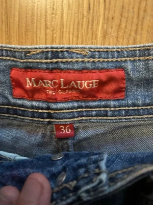 Blå raka jeans från Marc Lauge - Klassiska blå jeans från Marc Lauge med rak passform och normal midja. Jeansen har fem fickor och stängs med knappar. Perfekta till en avslappnad stil och tillverkade i slitstarkt denimtyg.