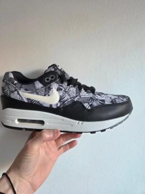 Nike Air Max 1 GPX Floral - Nike Air Max 1 sneakers i svart och vitt med ett unikt blommigt mönster på ovandelen. Skon har en vit sula med synlig Air-enhet, svart läderdetalj och vit Swoosh-logga på sidan. Klassisk låg siluett och svarta skosnören. Perfekt för dig som gillar statement sneakers.