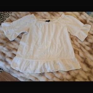 Vit Blus med Volang - Söt vit blus med broderade hål och volangkant nedtill. Blusen har trekvartsärm och resår vid axlarna för en avslappnad look. Perfekt till jeans eller kjol för en trendig outfit.