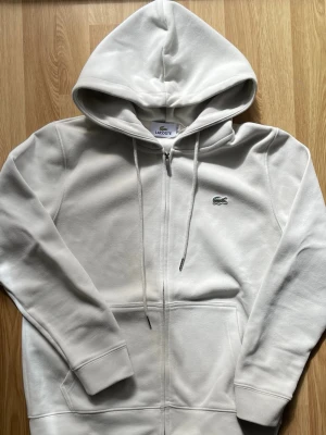 Vit zip hoodie från Lacoste - Säljer en clean vit hoodie från Lacoste med dragkedja, huva med dragsnören och klassiska krokodilloggan broderad på bröstet. Hoodien har två fickor framtill och är gjord i mjuk bomullsblandning som känns riktigt skön mot huden.