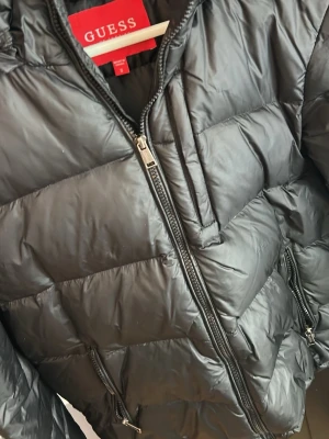 Svart pufferjacka från Guess - Snygg svart pufferjacka från Guess med dragkedja framtill och flera praktiska fickor. Jackan har en quiltad design och Guess-logga på ärmen. Tillverkad i glansigt syntetmaterial som håller värmen under kalla dagar. Perfekt för dig som vill ha en stilren vinterjacka.