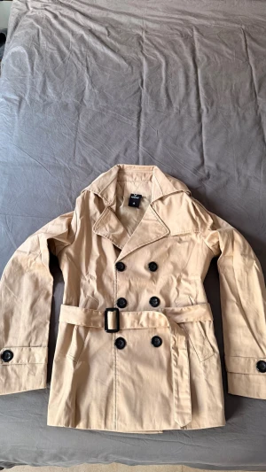 Beige trenchcoat med bälte och knappar. HELT NY - Helt ny stilren beige trenchcoat med dubbelknäppning, bred krage och bälte i midjan. Jackan har svarta knappar och klassiska detaljer som axelklaffar och slejf vid ärmslut. Perfekt för lager-på-lager och snygg till alla outfits.