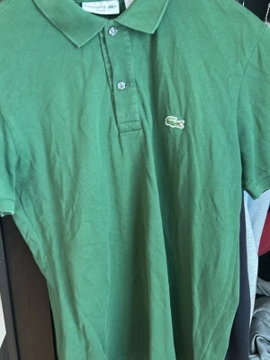 Grön pikétröja från Lacoste - Säljer en grön pikétröja från Lacoste i slim fit-modell. Tröjan har klassisk krage, korta ärmar och knappar framtill. Ikoniska krokodilloggan är broderad på bröstet. Tillverkad i bomull för en skön känsla.