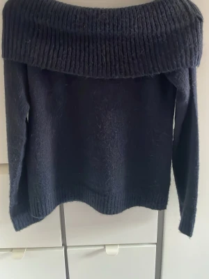 Marinblå offshoulder stickad tröja Gina Tricot - Mysig svart stickad tröja från Gina Tricot med bred ribbad krage som sitter offshoulder. Tröjan har lång ärm och är perfekt för dig som gillar en chill och snygg look. Materialet är mjukt och värmande, passar dig som vill ha något stilrent och enkelt.