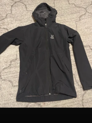 Svart vindjacka från Haglöfs - Snygg svart vindjacka från Haglöfs med huva och hel dragkedja framtill. Jackan har två snedställda fickor med dragkedja och meshfoder på insidan för extra ventilation. Perfekt för blåsiga dagar och outdoor-aktiviteter.                                                        (Nypris: 1799kr❌ vs  Mitt pris 1000kr✅)