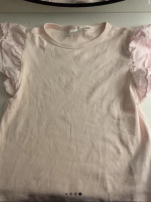 Ljusrosa topp med volangärmar - Supersöt ljusrosa topp i ribbad bomull med korta volangärmar i spets. Perfekt för dig som gillar romantiska detaljer och en feminin look. Toppen har rund halsringning och är lätt att matcha med både jeans och kjol.