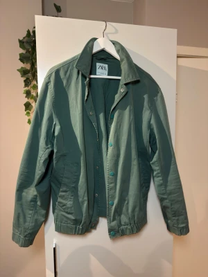 Grön casual jacka från Zara - Stilren grön jacka från Zara med klassisk krage och tryckknappar framtill. Jackan har två sidofickor och är tillverkad i ett mjukt bomullsliknande material. Perfekt för dig som vill ha en enkel men snygg jacka till vardags.