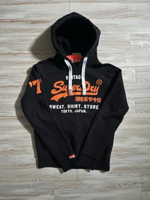 Svart Superdry hoodie med tryck - Svart hoodie från Superdry med stor orange logga och text på bröstet samt vitt tryck🙌🏻 Snygg detalj med orange '77 på ärmen🤩 Inga defekter som jag vet om, skriv gärna vid funderingar.