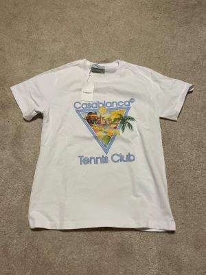 Vit Casablanca Tennis Club t-shirt - Säljer en vit t-shirt från Casablanca med färgglatt tryck och texten 'Tennis Club' på bröstet. T-shirten har klassisk passform, korta ärmar och är gjord i mjuk bomull. Perfekt för dig som gillar sportig och trendig stil.