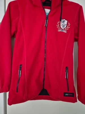 Fleecejacka - Röd fleecejacka med huva. Jackan har dragkedja framtill, två fickor med dragkedja och broderad logga på bröstet. Mjuk och skön fleece, perfekt för kyliga dagar. Elastiska muddar vid ärmsluten och längre passform baktill. Har några fläckar.