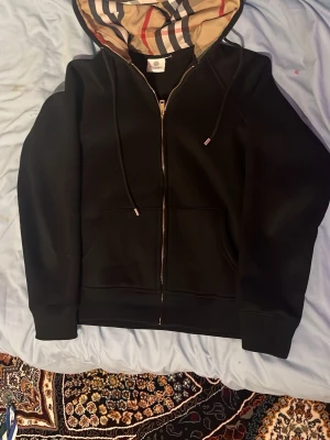 Svart zip hoodie från Burberry - Snygg svart hoodie med dragkedja från Burberry. Klassisk huva med det ikoniska.          De är rep. rutmönstret på insidan och justerbara snören. Mjukt material och ribbade muddar vid ärmar och nederkant. Perfekt för en stilren och avslappnad look. Den passar dig som är 169 til 172 cm. Och priset kan diskuteras tveka inte att skicka ett meddelande 