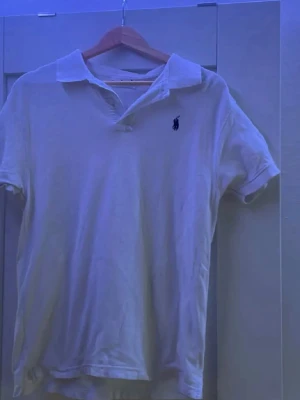 Vit piké från Ralph Lauren - Klassisk vit pikétröja från Ralph Lauren med korta ärmar och den ikoniska loggan broderad på bröstet. Tröjan har krage och knappar vid halsen. Perfekt till en avslappnad men ändå stilren look.