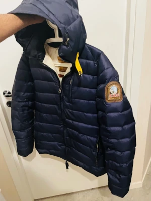 Mörkblå pufferjacka från Parajumpers - Snygg mörkblå pufferjacka från Parajumpers med huva och gul detalj vid halsen. Jackan har patch med logga på ärmen, dragkedja framtill och quiltad design. Perfekt för kalla vinterdagar och riktigt varm. Materialet är glansigt och jackan har flera praktiska fickor.