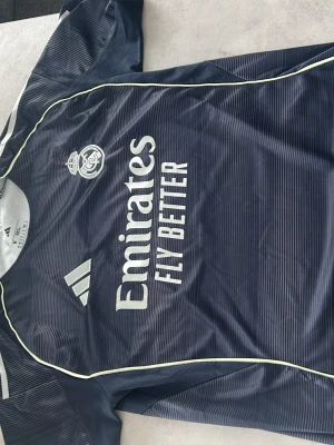 Real Madrid tränings t-shirt Adidas - Mörkblå Real Madrid tränings t-shirt från Adidas med tunna vita ränder, klubbmärke och Adidas-logga på bröstet samt 'Emirates Fly Better' tryckt framtill. Tillverkad i lätt och ventilerande funktionsmaterial, perfekt för fotbollsträning.