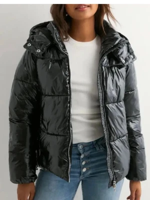 Svart glansig pufferjacka med huva - Snygg svart pufferjacka med glansig finish och stor huva. Jackan har breda kanaler, dragkedja framtill och tryckknappar vid kragen. Perfekt för kalla dagar när du vill hålla stilen och värmen.