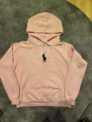 Ljusrosa hoodie från Polo Ralph Lauren - Snygg ljusrosa hoodie från Polo Ralph Lauren med stor mörkblå logga på bröstet. Hoodien har huva med vita dragsnören och en klassisk magficka. Tillverkad i mjukt bomullsmaterial och har ribbade muddar vid ärmar och nederkant. Tröjan står att det är storlek XL men den är egentligen storlek S-M, jag är 183 och tröjan sitter som på bilden på mig.